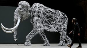 Ein lebensgroßes Mammut aus weißen Kabeln steht in einer Halle; ein Mann geht daneben her und hält ein Kabel in der Hand, das zu der Figur führt.