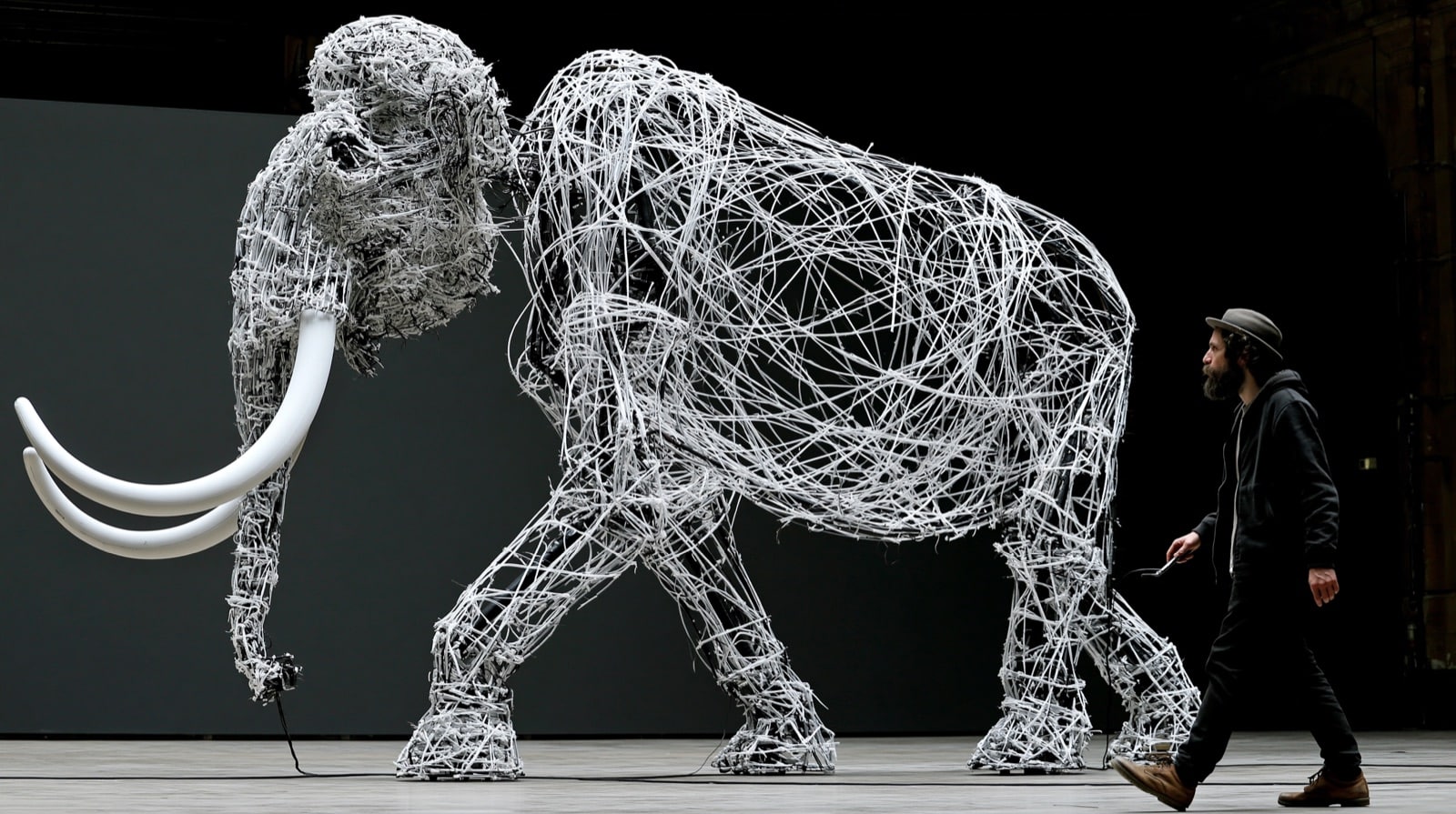 Projektarchäologie: Warum abgebrochene Vorhaben eine zweite Bewertung verdienen 1 Ein lebensgroßes Mammut aus weißen Kabeln steht in einer Halle; ein Mann geht daneben her und hält ein Kabel in der Hand, das zu der Figur führt.